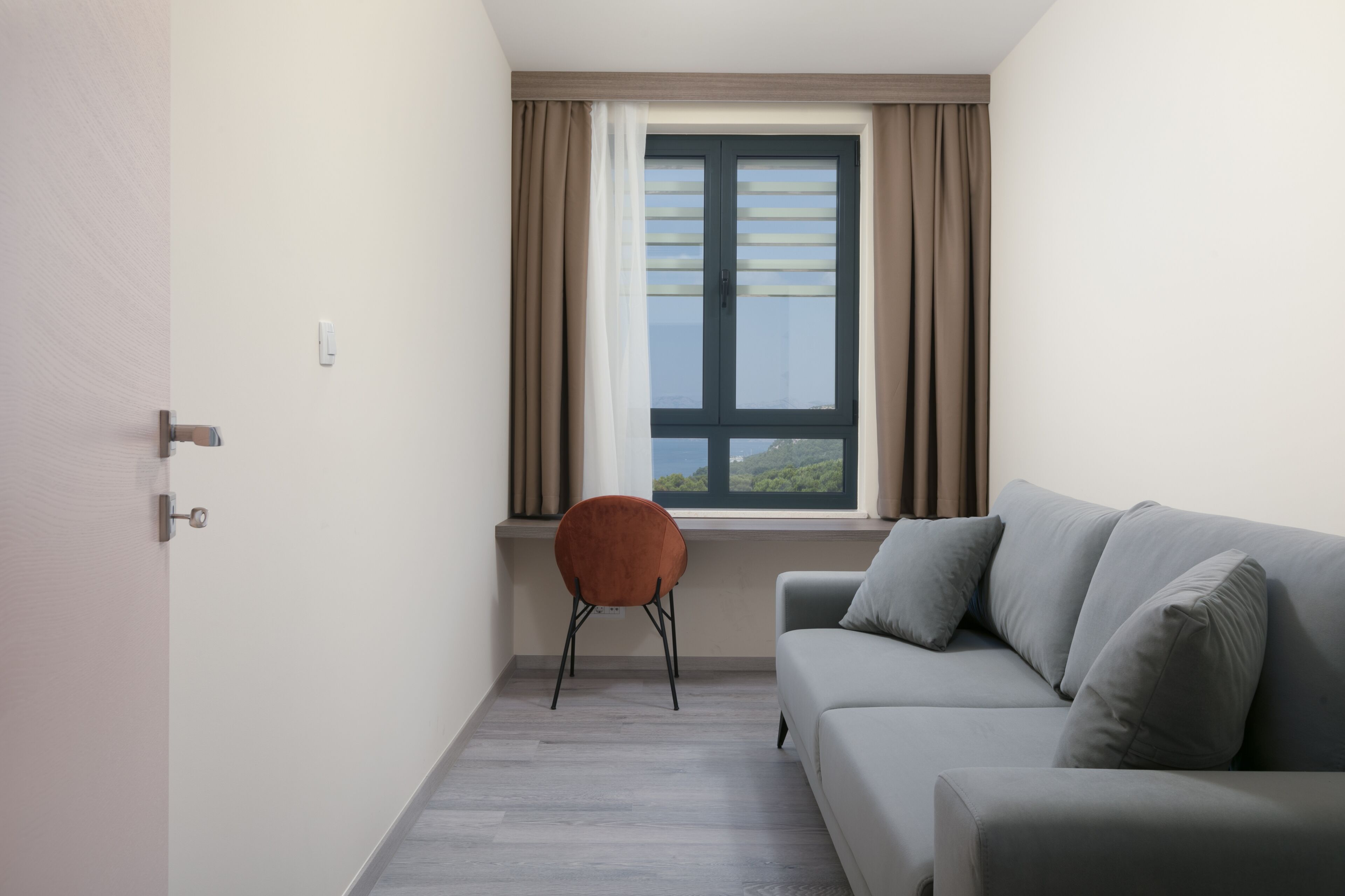 Un salon moderne avec un canapé gris, un fauteuil marron, une fenêtre avec des rideaux et une porte.