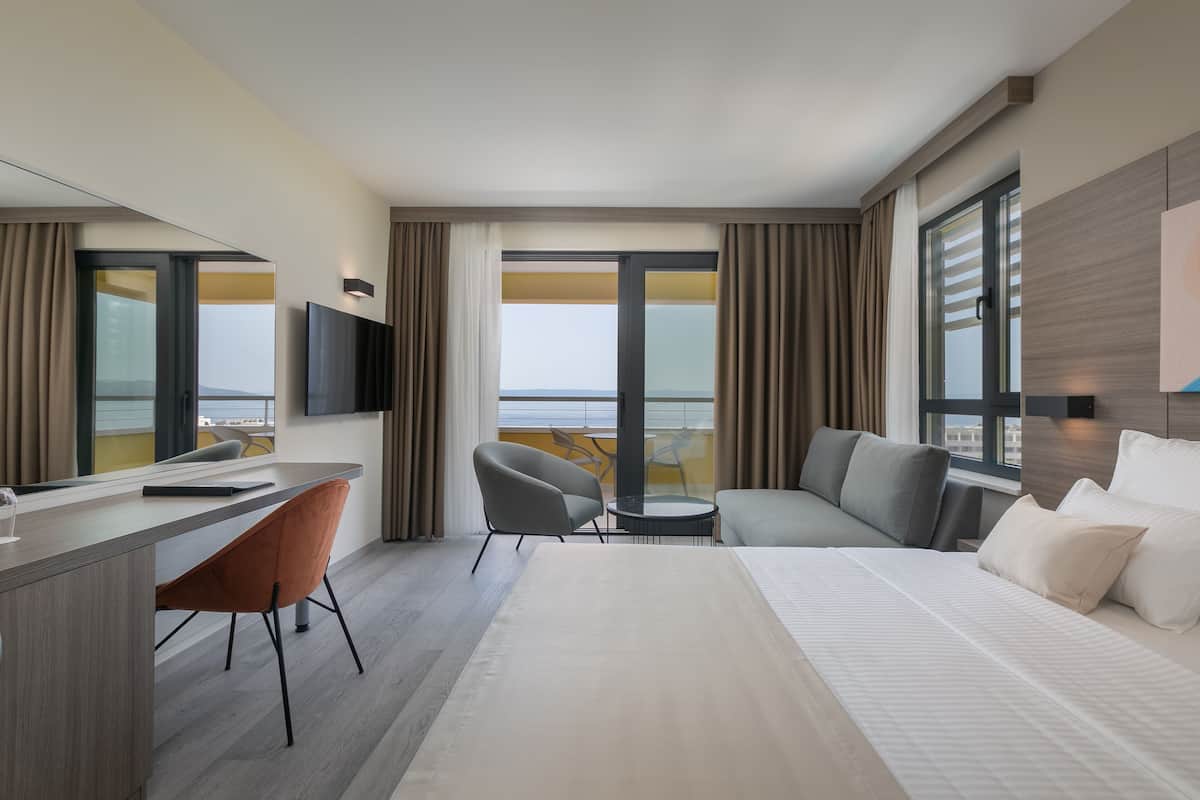 A modern hotel room with a large bed, a desk, a chair, a sofa, and a TV.
