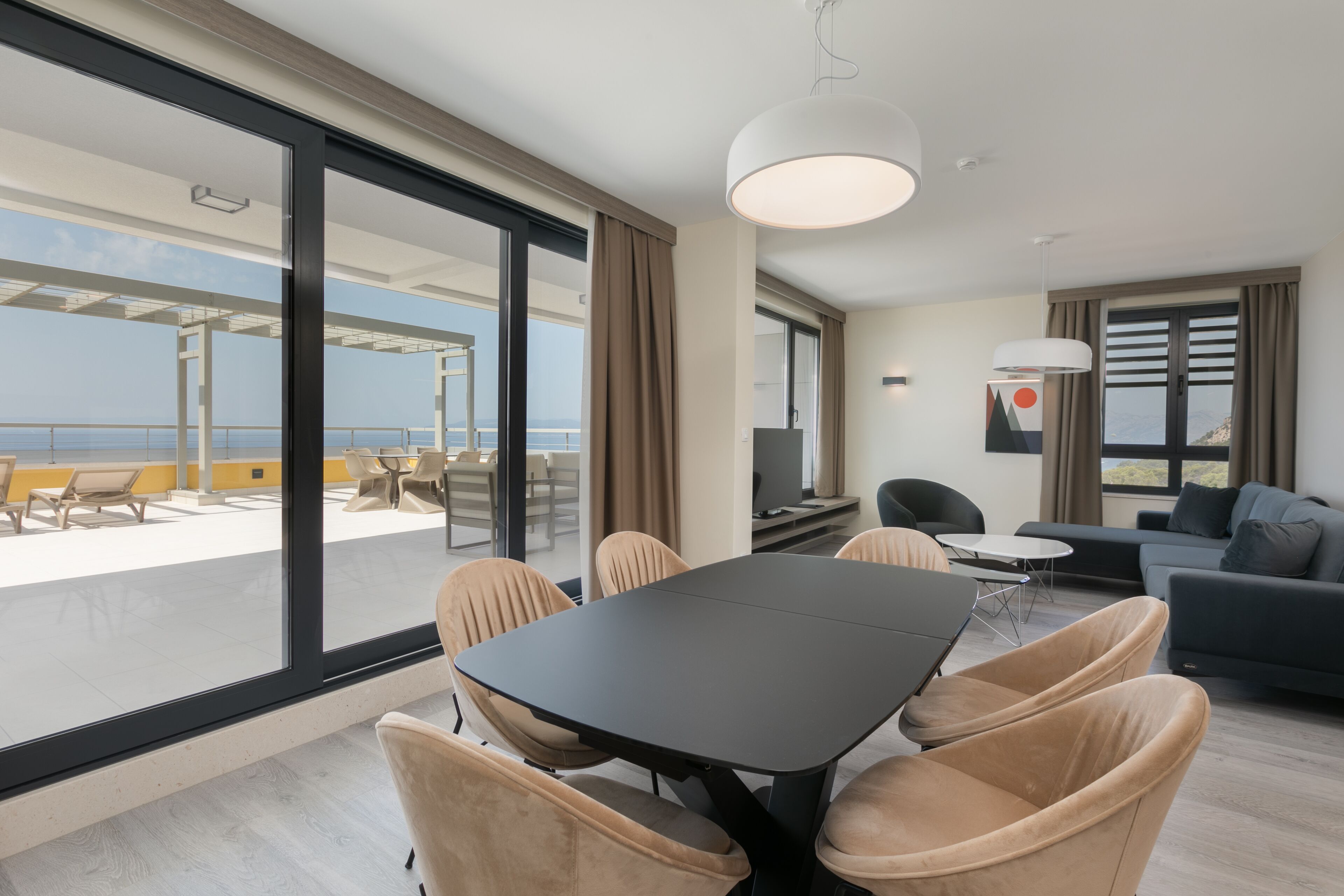 A modern hotel room with a large balcony, a dining area with a table and chairs, a sofa, and a television.