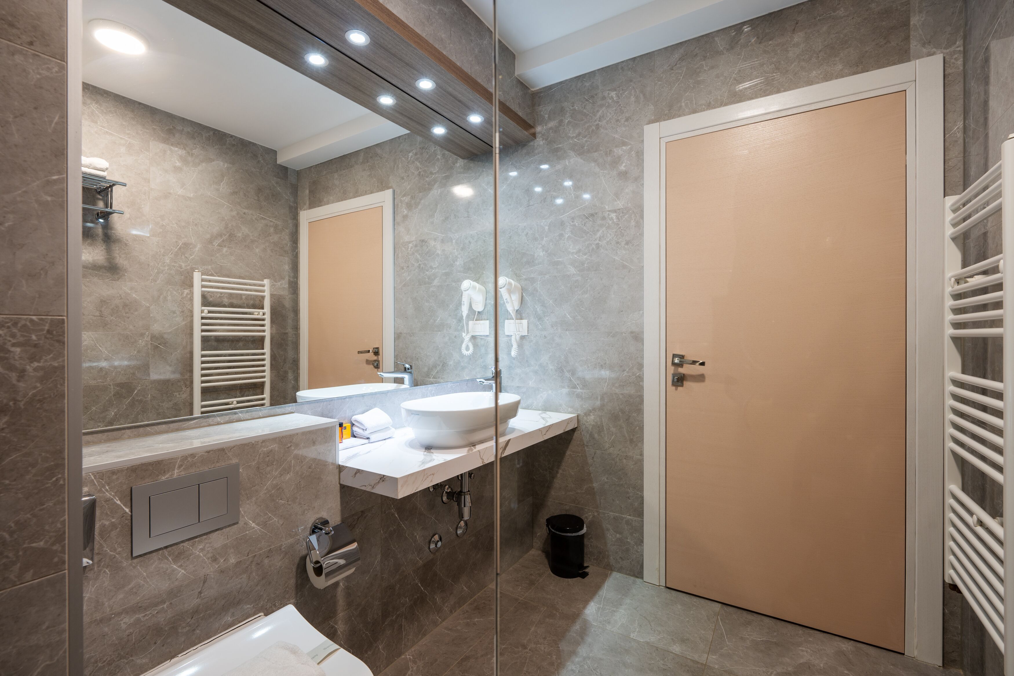 A modern bathroom with a marble wall, a white sink, a large mirror, and a beige door.