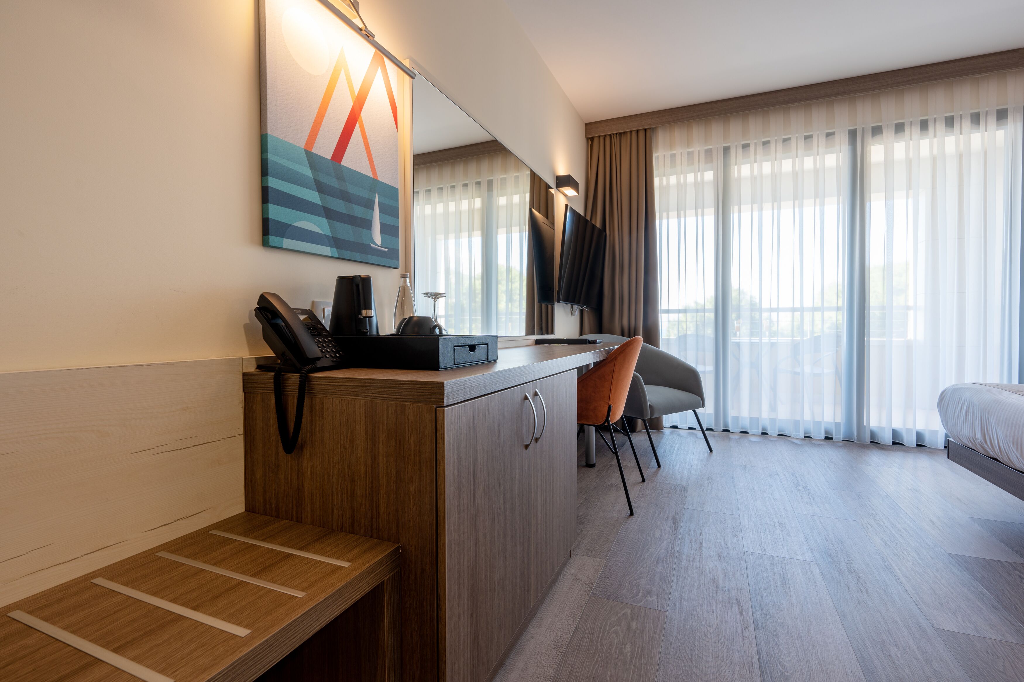 Habitación de hotel con escritorio de madera, una silla, una cama, un televisor y un ventanal con cortinas.