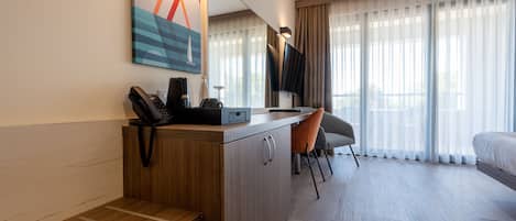 Habitación de hotel con escritorio de madera, una silla, una cama, un televisor y un ventanal con cortinas.