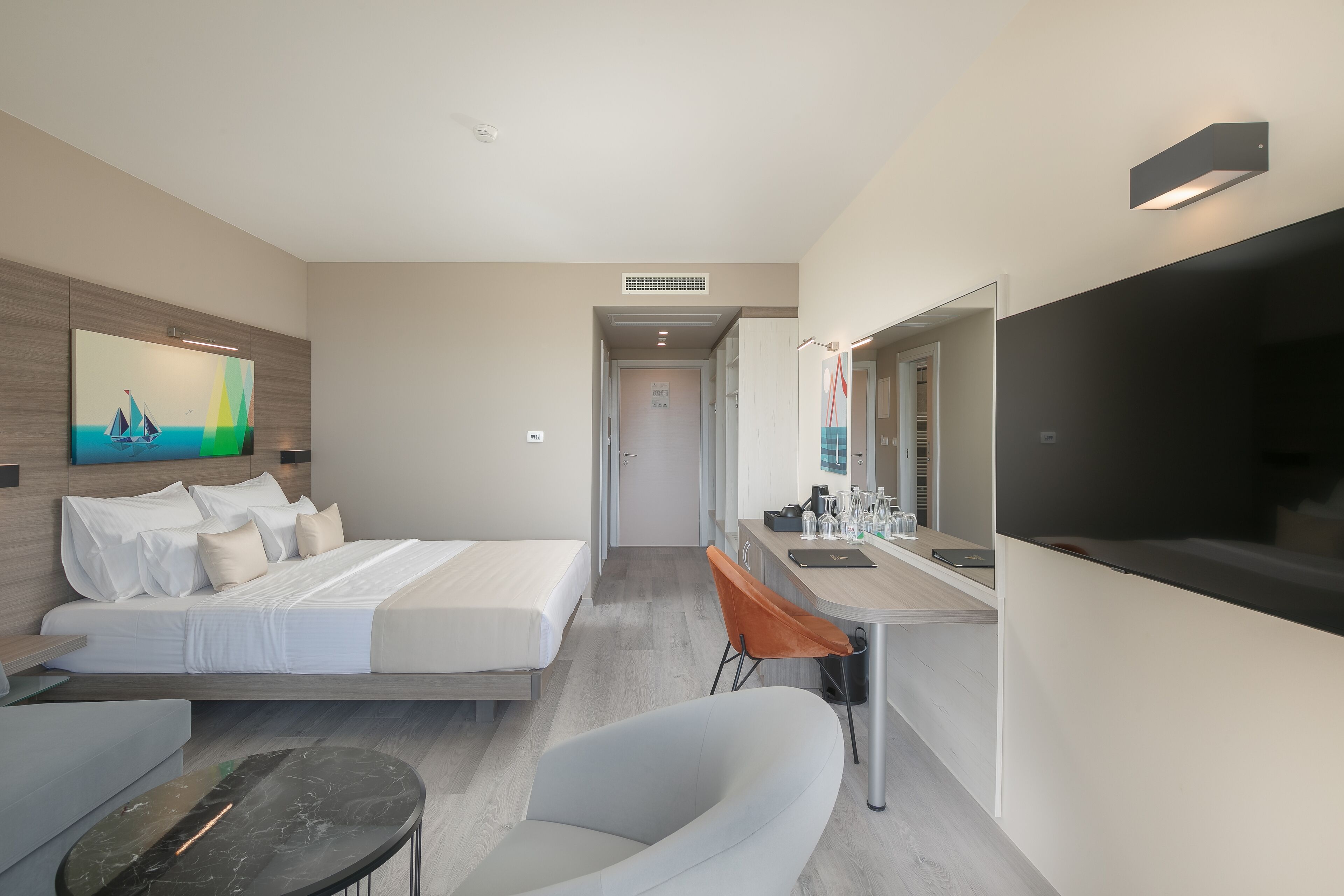A modern hotel room with a bed, a sofa, a desk with a mirror, and a TV.