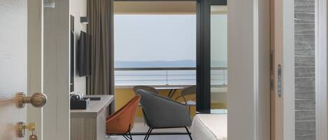 A hotel room with a bed, a desk, a chair, and a TV. There is a view of the sea through large windows.