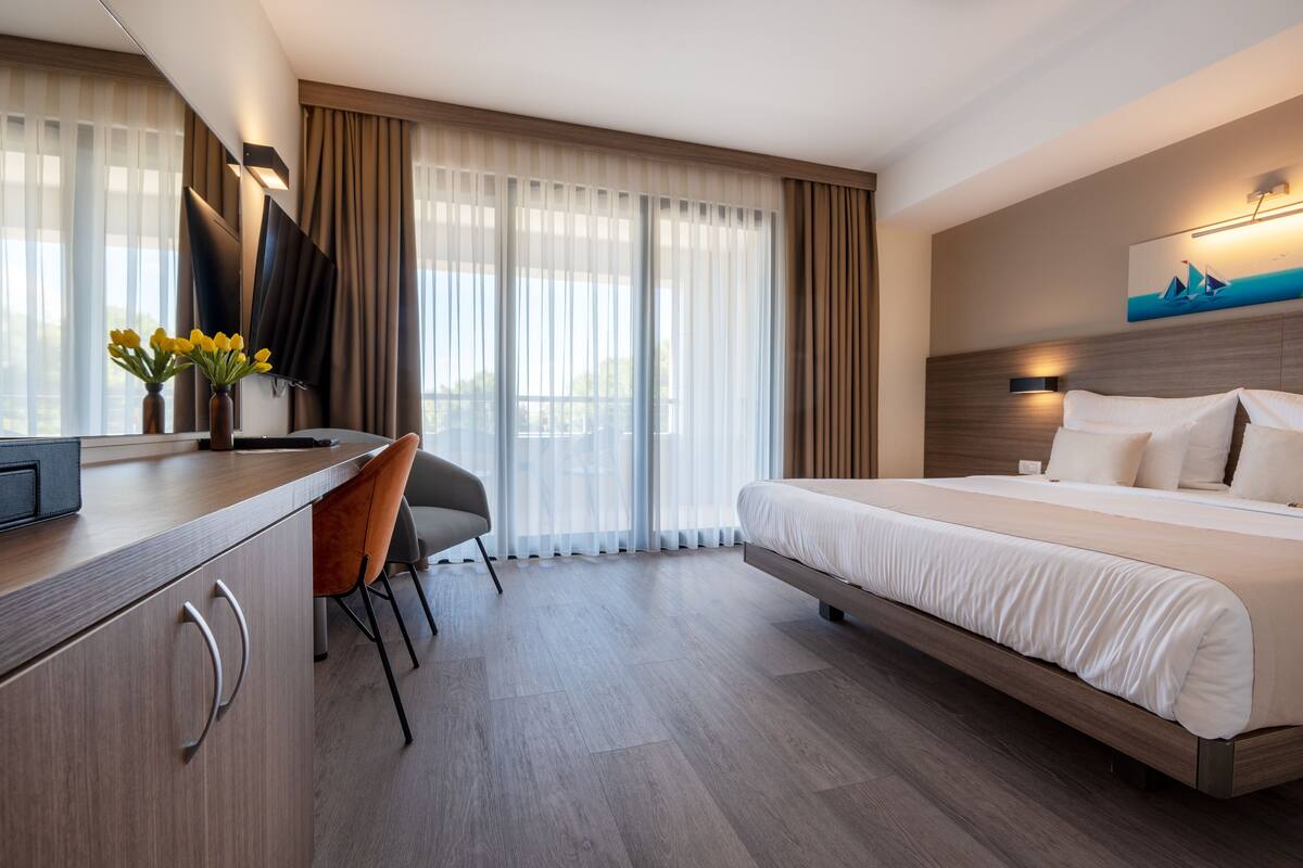 A modern hotel room with a bed, a desk, a chair, and a large window with curtains.