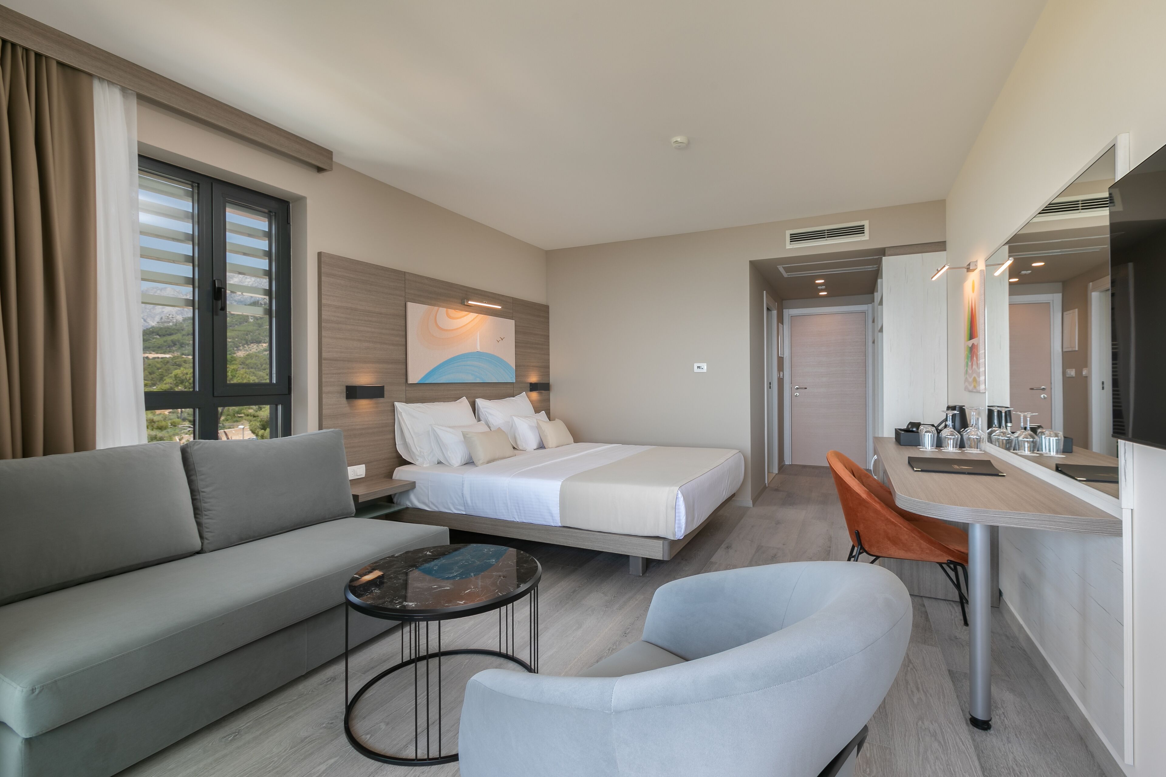 A modern hotel room with a bed, sofa, dining table, and a small seating area.
