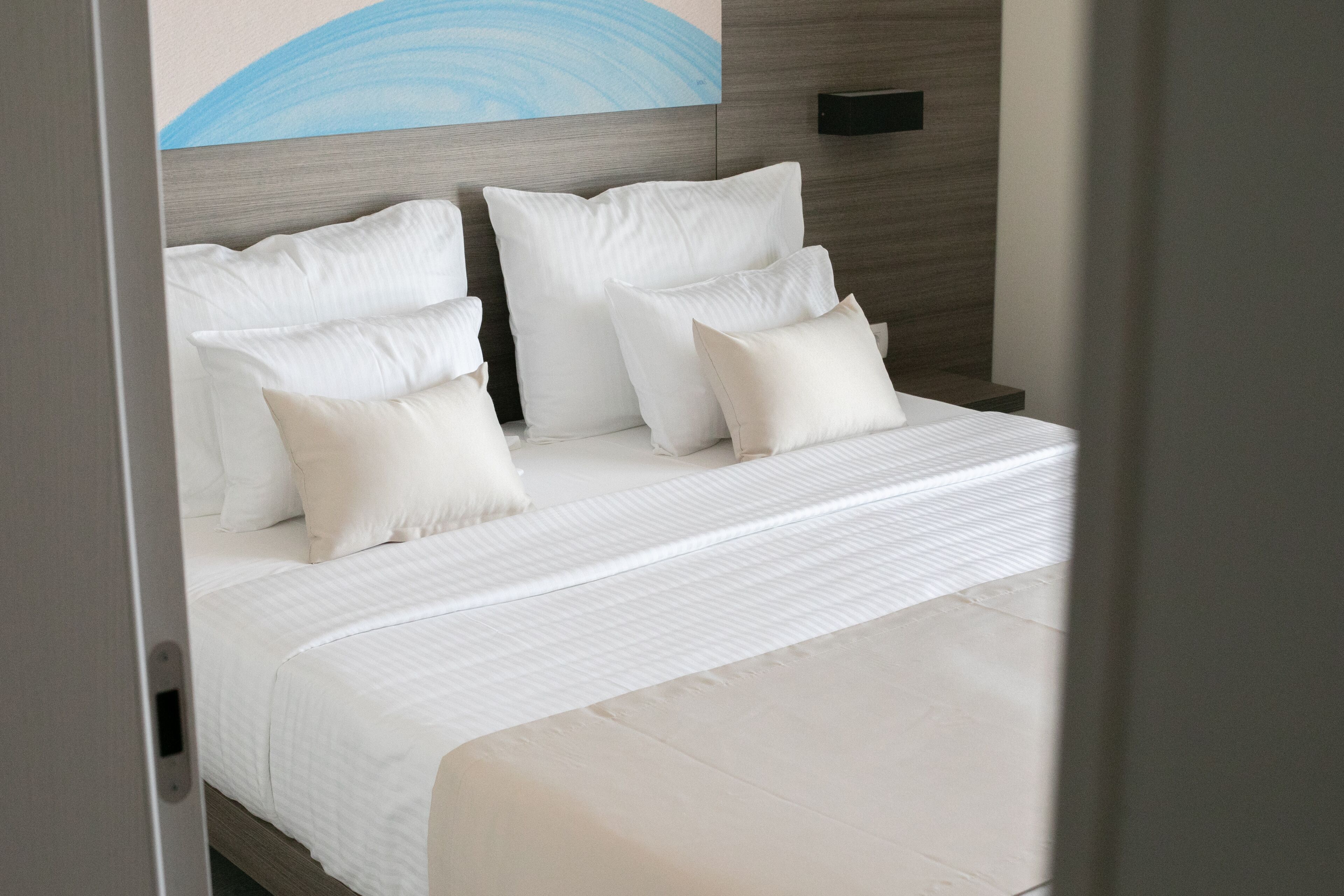 A neatly made bed with white linens and pillows, a wooden headboard, and a wall-mounted light fixture.