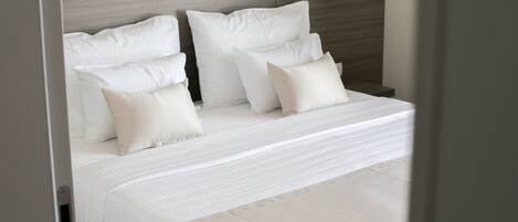 Una cama bien hecha con sábanas y almohadas blancas, cabecera de madera y lámpara de pared.
