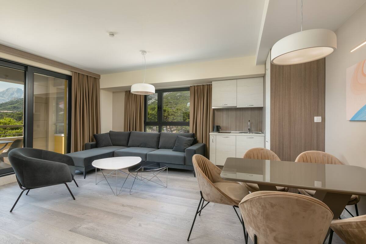 A modern living room with a gray sofa, a glass dining table, and beige chairs. There is a kitchen area with cabinets and a window.