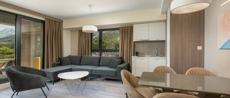 A modern living room with a gray sofa, a glass dining table, and beige chairs. There is a kitchen area with cabinets and a window.