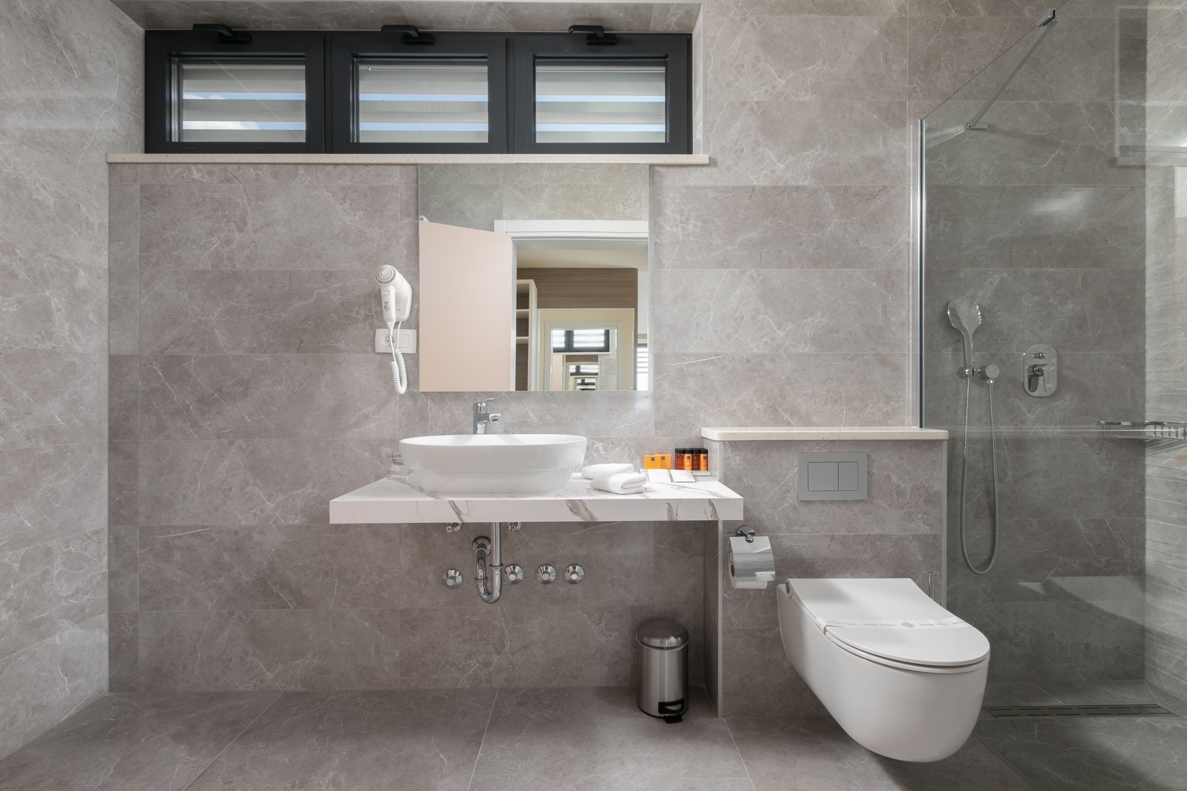 A modern bathroom with a white sink, mirror, and toilet, a glass-enclosed shower, and a trash can.