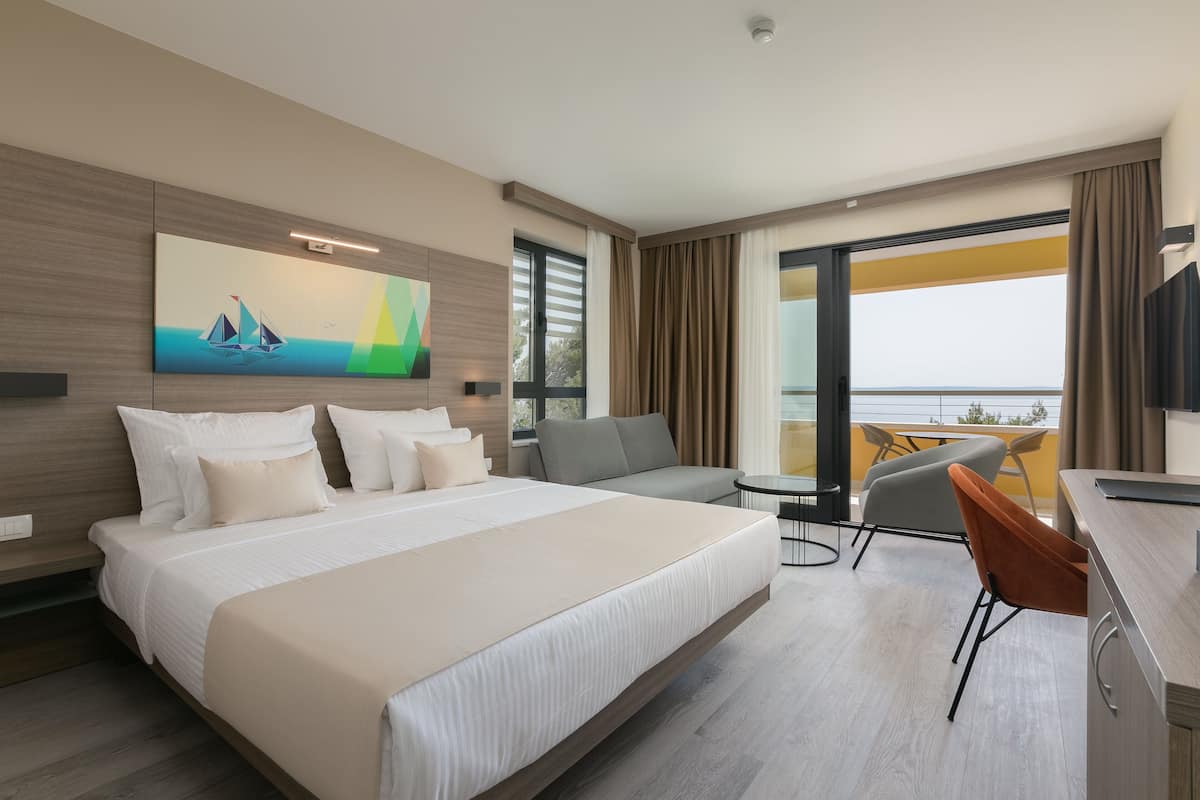 A modern hotel room with a large bed, a sofa, a desk, and a balcony with a view.
