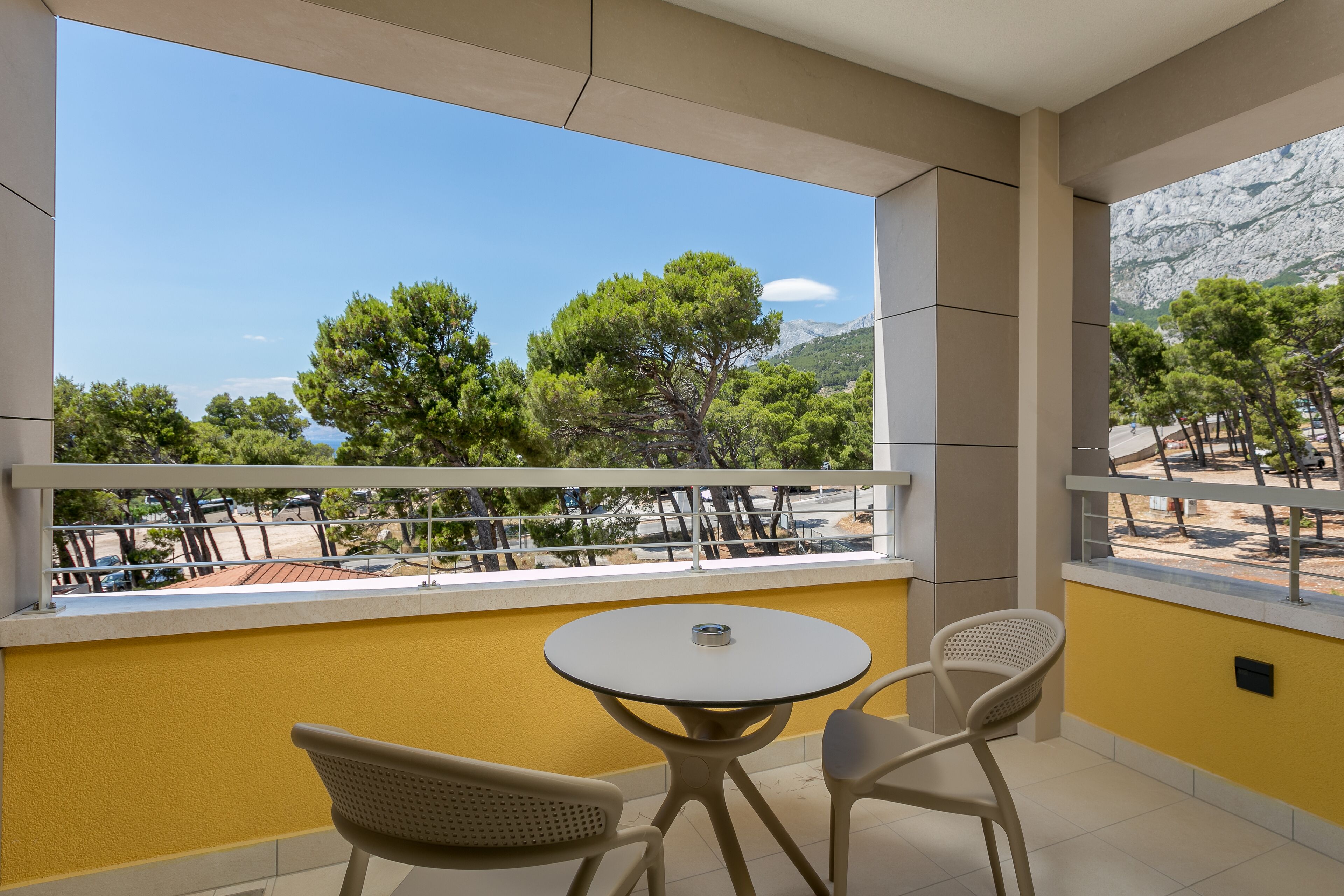 A balcony with a yellow wall, two chairs, a small round table, and a view of trees and mountains.