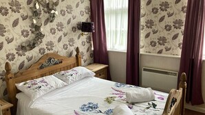 A bedroom with a wooden bed, floral bedding, a bedside table, a wall-mounted lamp, and a window with purple curtains. - The Tayleur Arms (Telford)