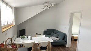 Un salon moderne comprenant un coin repas, un canapé et un téléviseur. On y trouve une fenêtre avec une étagère en bois, un panier rempli de fruits et un luminaire suspendu au plafond.