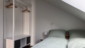 Une chambre avec un plafond en pente, une armoire blanche avec des cintres en bois, une table de chevet avec une lampe et un lit avec une literie verte.