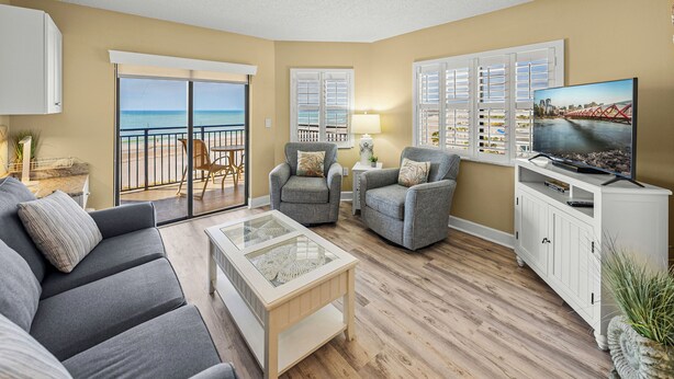 A living room with a sofa, armchairs, a coffee table, and a TV on a stand. There is a balcony with a view of the beach.