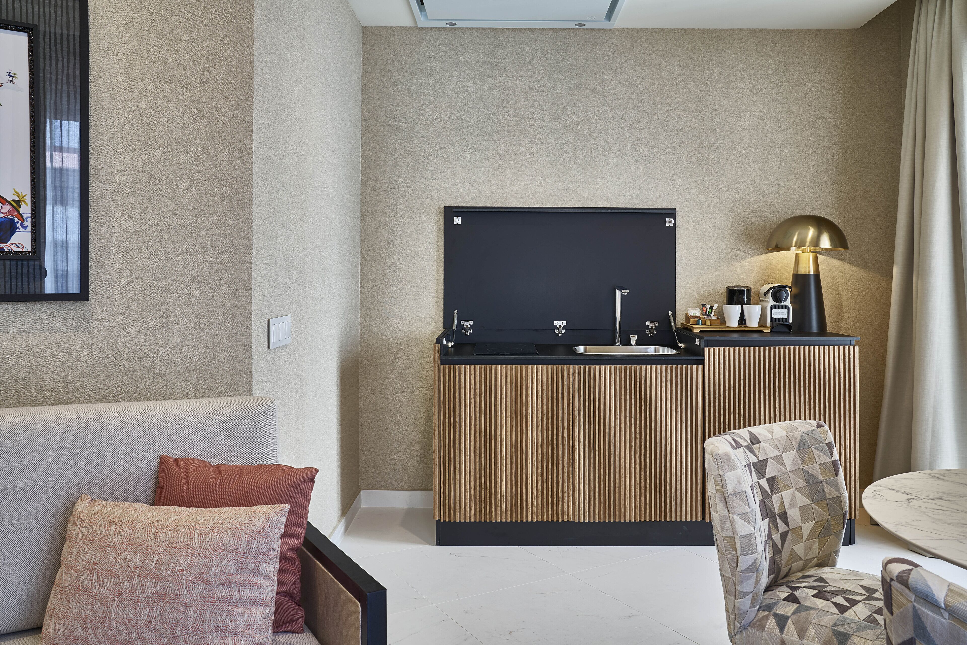 A modern hotel room with a sofa, a patterned armchair, a small round table, a kitchenette with a blackboard, and a lamp.