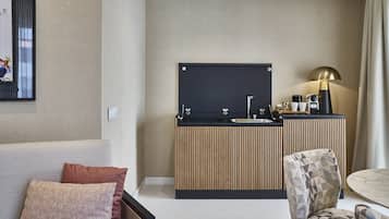 A modern hotel room with a sofa, a patterned armchair, a small round table, a kitchenette with a blackboard, and a lamp.