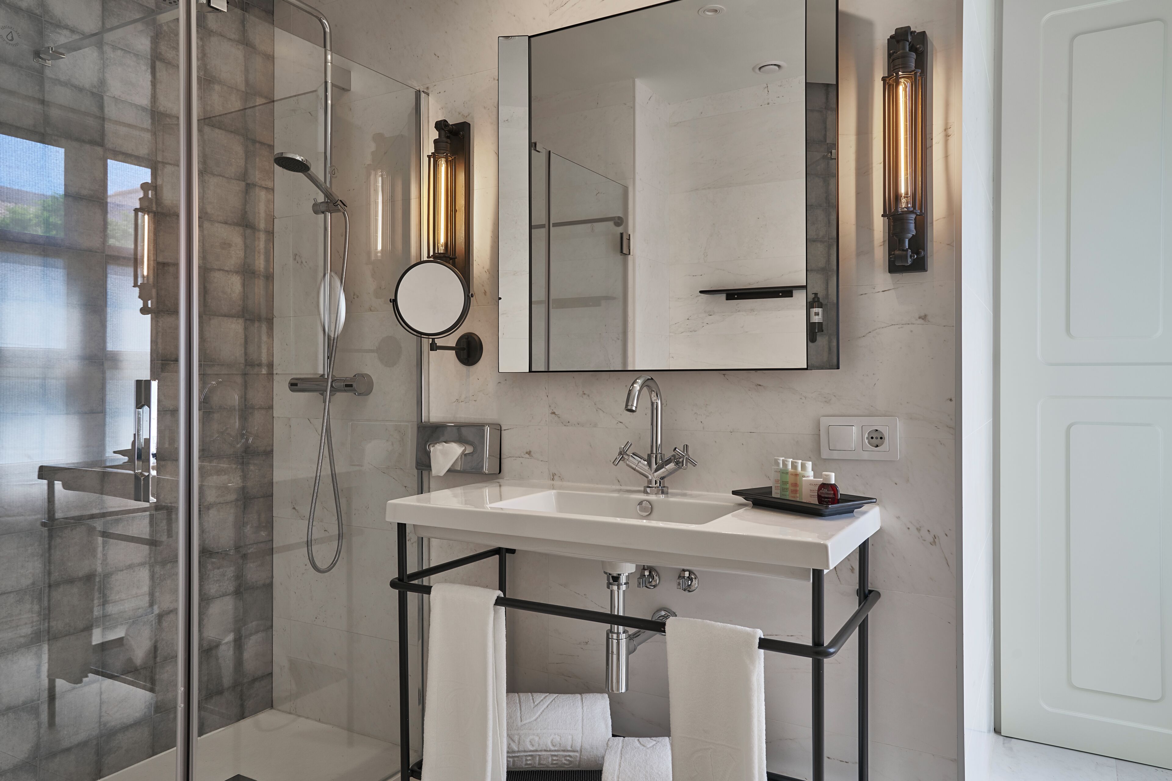 Une salle de bain moderne avec une douche à l’italienne en verre, un lavabo blanc avec un porte-serviettes noir et un grand miroir au-dessus du lavabo.