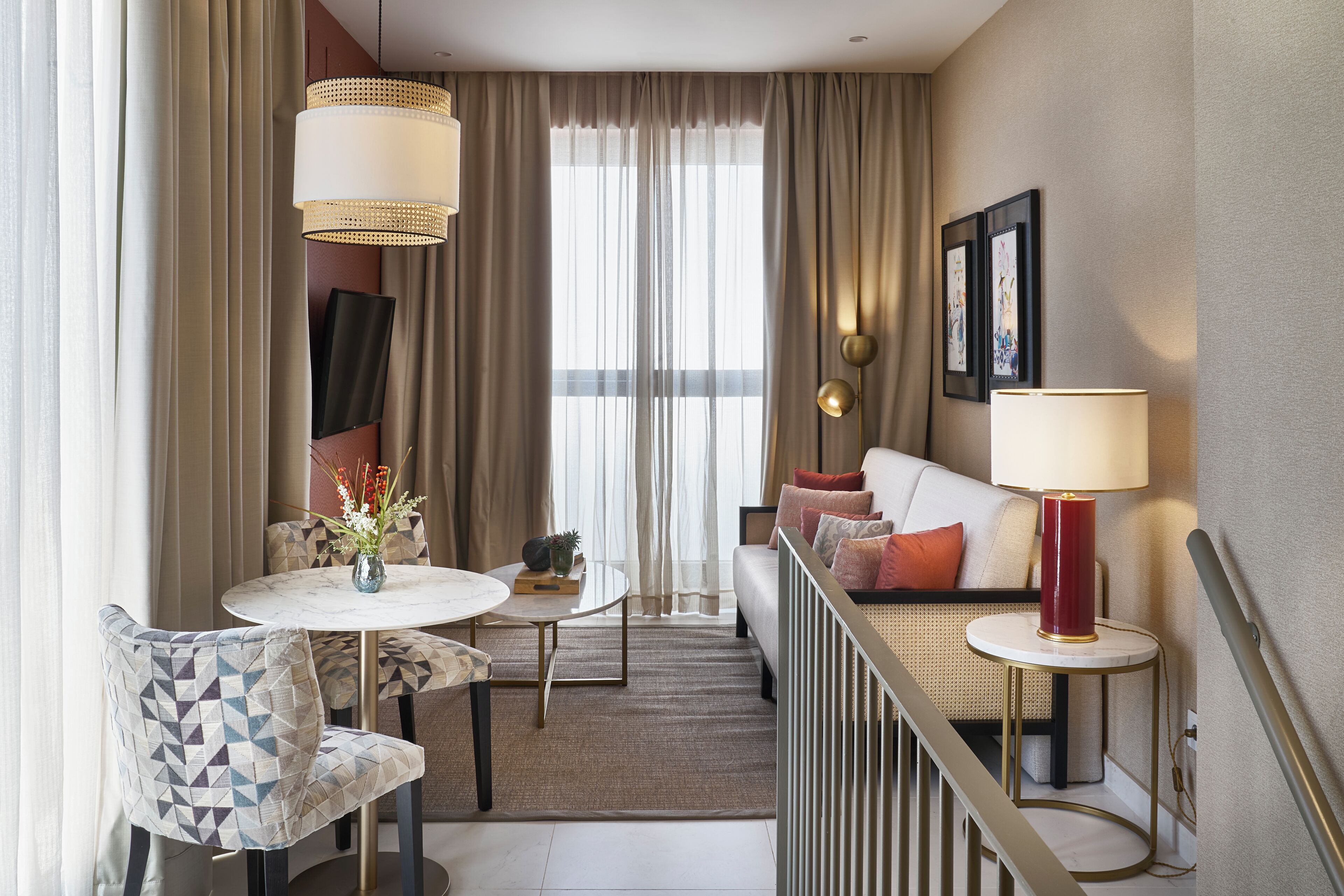 A hotel room with a dining area, a sofa with cushions, a red lamp, and a view through the curtains.