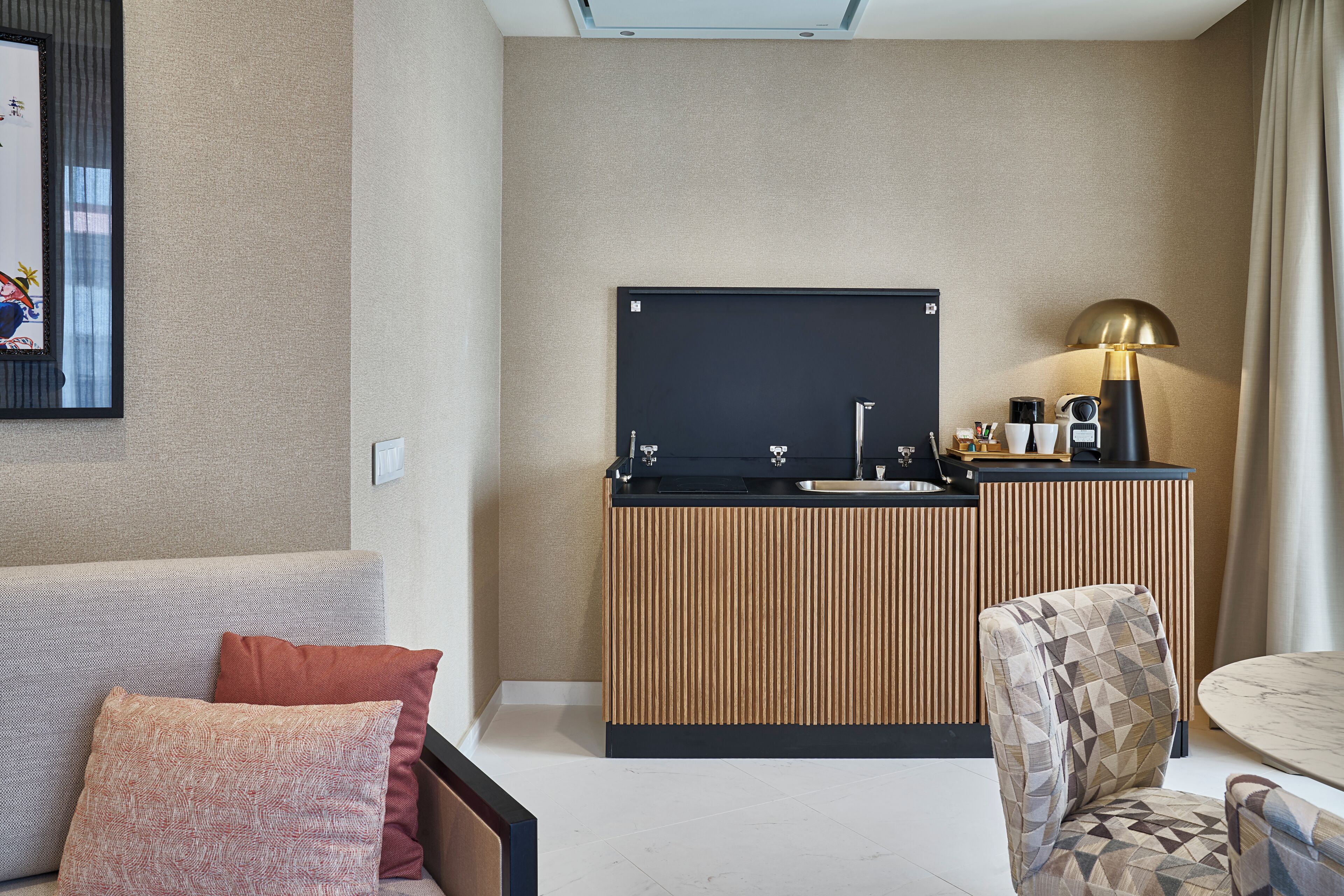 A modern hotel room with a sofa, a patterned armchair, a small round table, a kitchenette with a black countertop, and a mounted flat-screen TV.