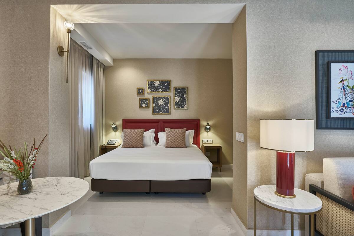 A hotel room with a large bed, a round table, a red lamp, and framed artwork on the wall.