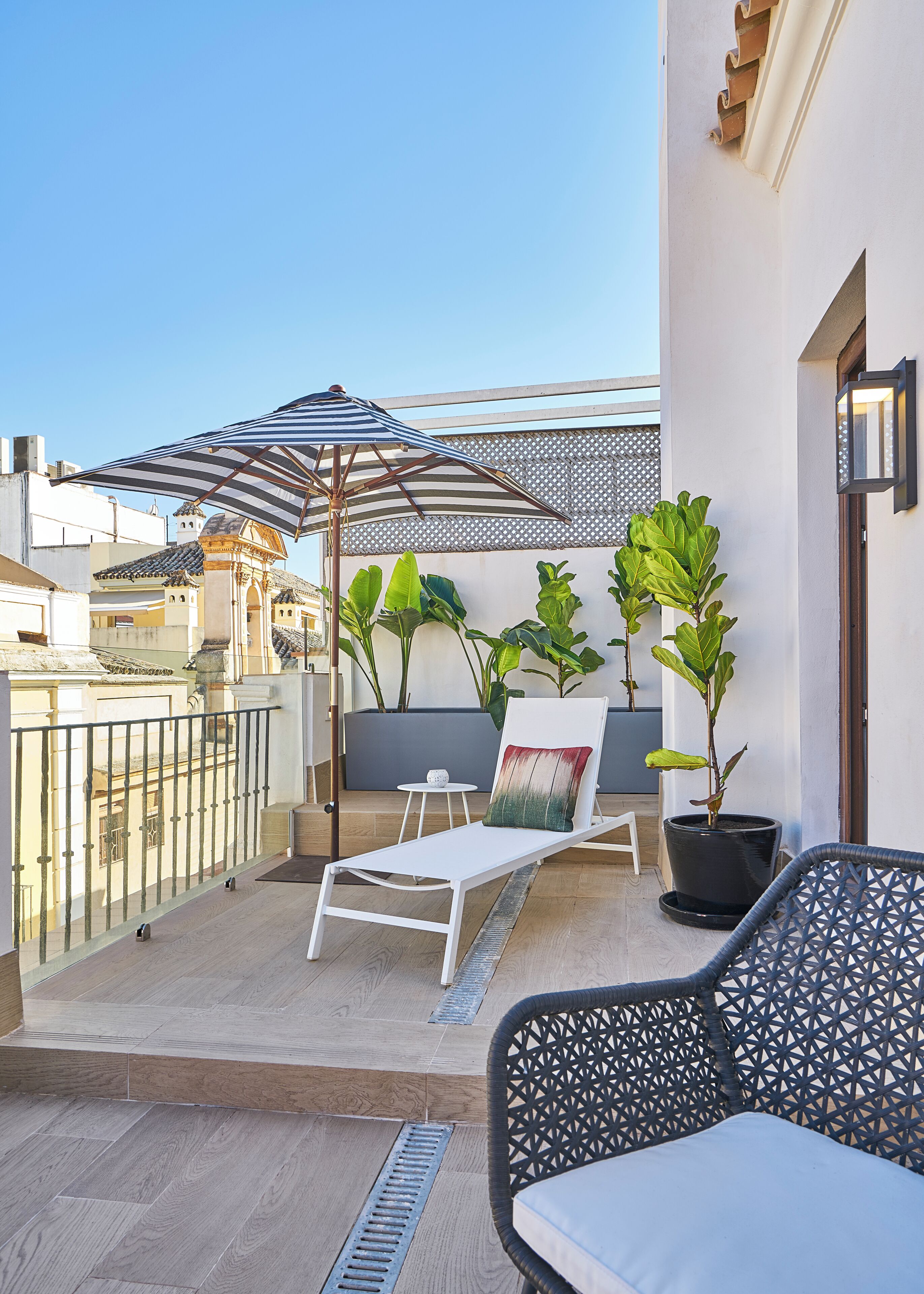 A rooftop terrace with a white lounge chair, a patterned chair, potted plants, a striped umbrella, and a view of the cityscape.