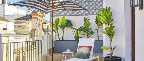 A rooftop terrace with a white lounge chair, a patterned chair, potted plants, a striped umbrella, and a view of the cityscape.
