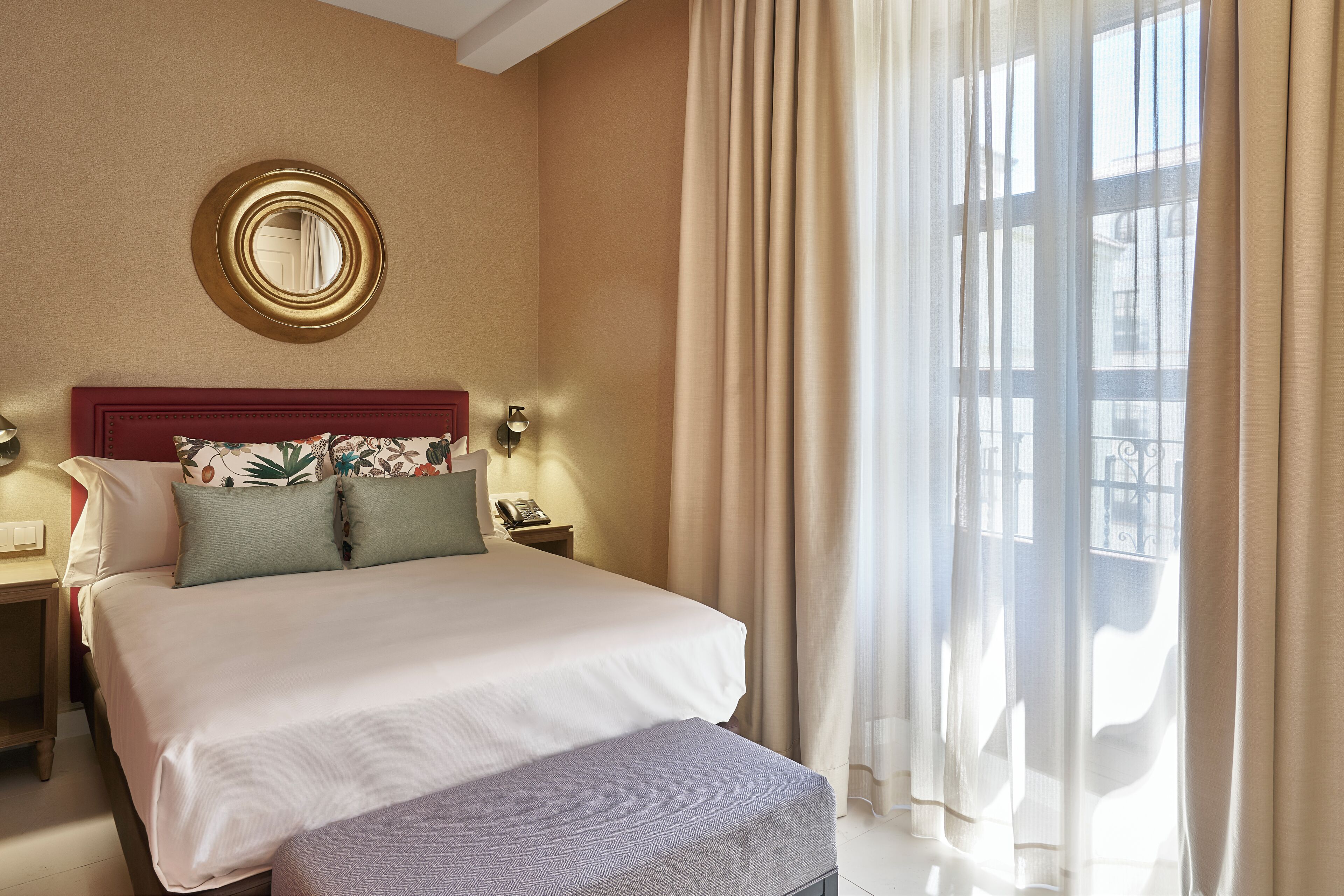 Een hotelkamer met een groot bed, een ronde spiegel, een nachtkastje met een telefoon en een raam met gordijnen.