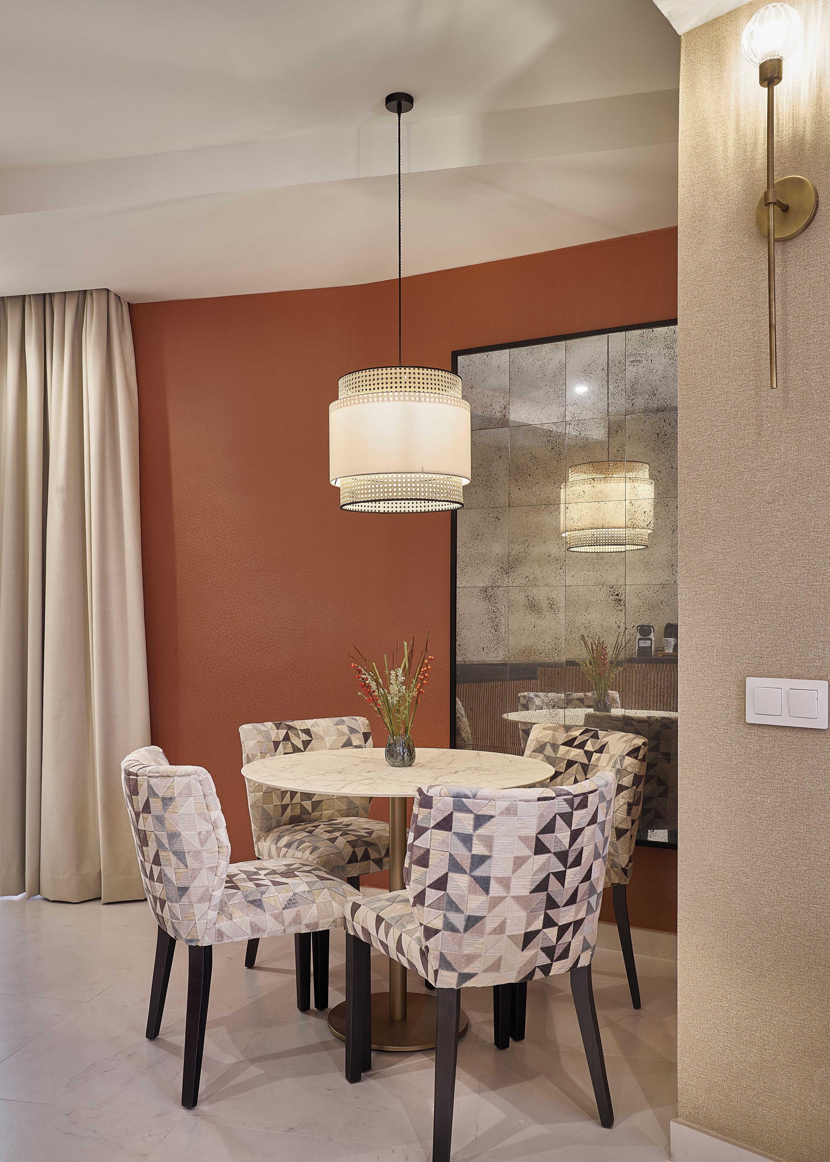 A dining area with a round table, patterned chairs, a hanging light fixture, and a vase with flowers.
