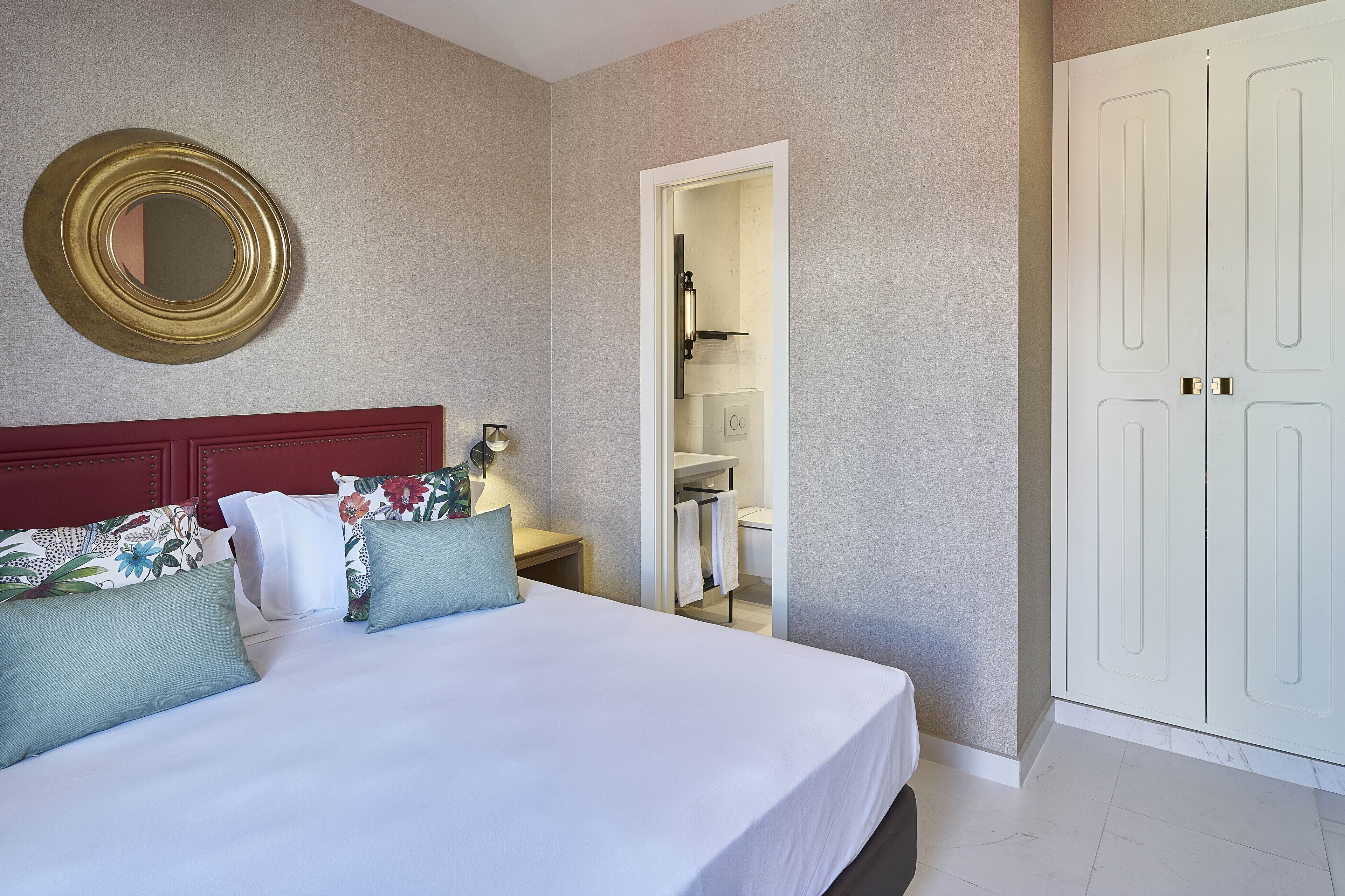 Una habitación de hotel con una cama grande, un cabecero rojo, un espejo redondo y un armario blanco.