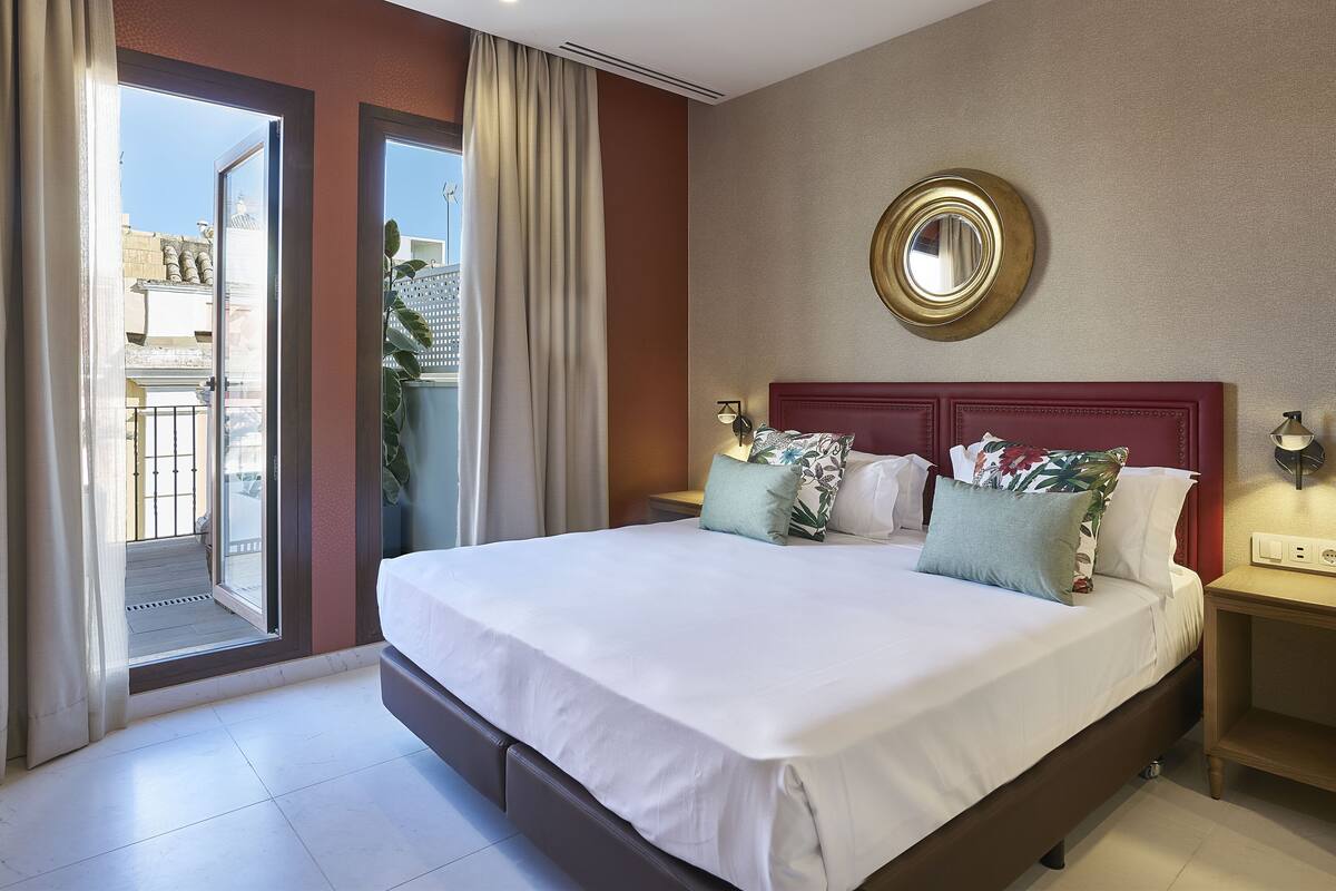 A hotel room with a large bed, a balcony with curtains, a round mirror on the wall, and a nightstand with a lamp.