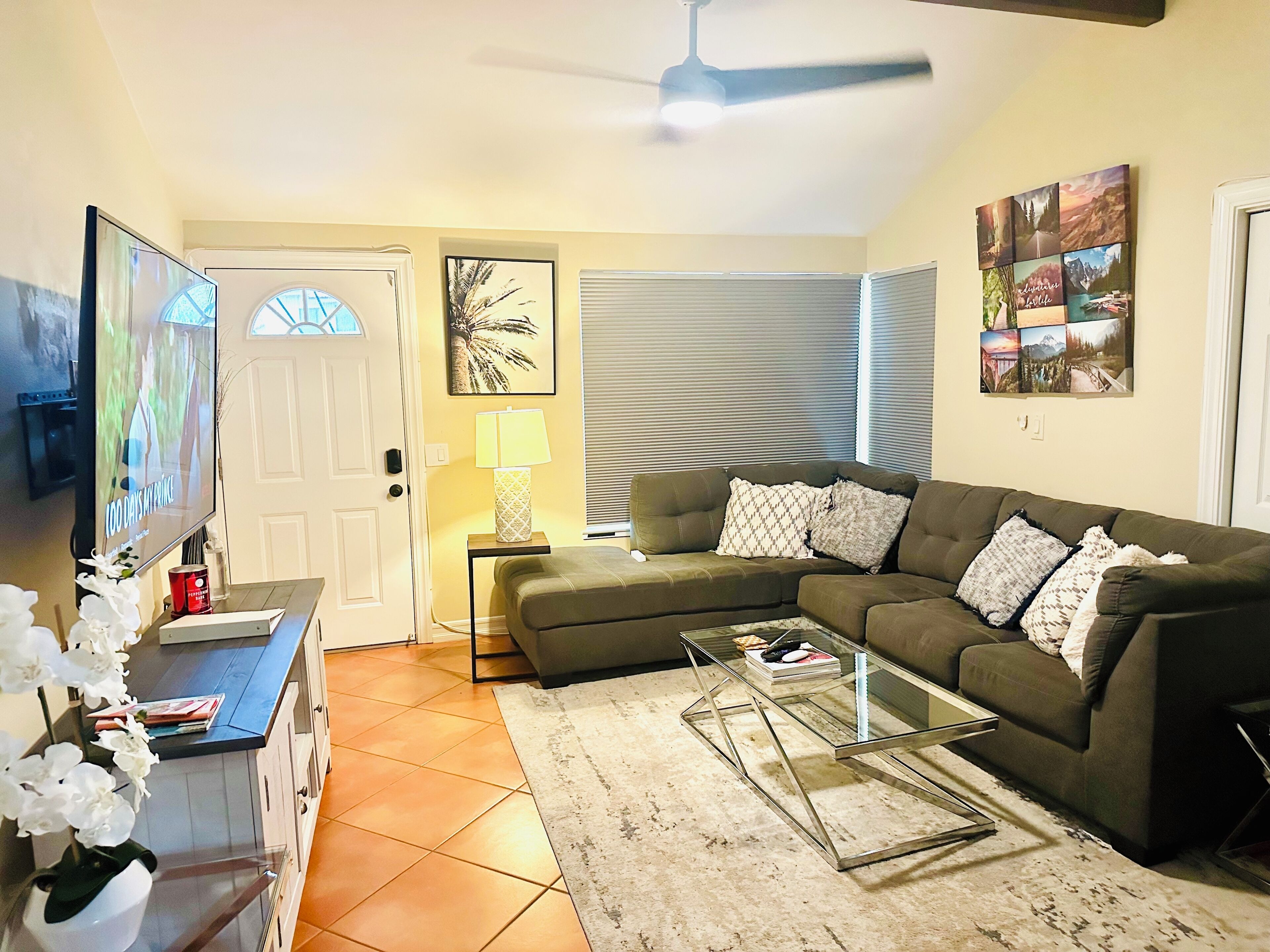 A living room with a sectional sofa, a glass coffee table, a TV, a lamp, and a door with blinds.