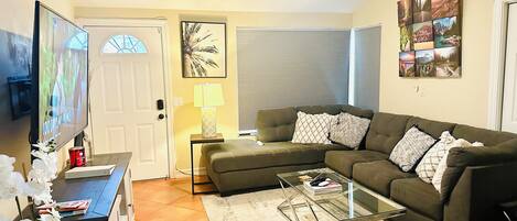 A living room with a sectional sofa, a glass coffee table, a TV, a lamp, and a door with blinds.