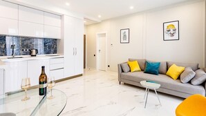 Una sala de estar moderna con un sofá, una mesa de centro de vidrio y una cocina con armarios blancos y un salpicadero de mármol.