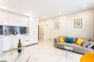 Una sala de estar moderna con un sofá, una mesa de centro de vidrio y una cocina con armarios blancos y un salpicadero de mármol.