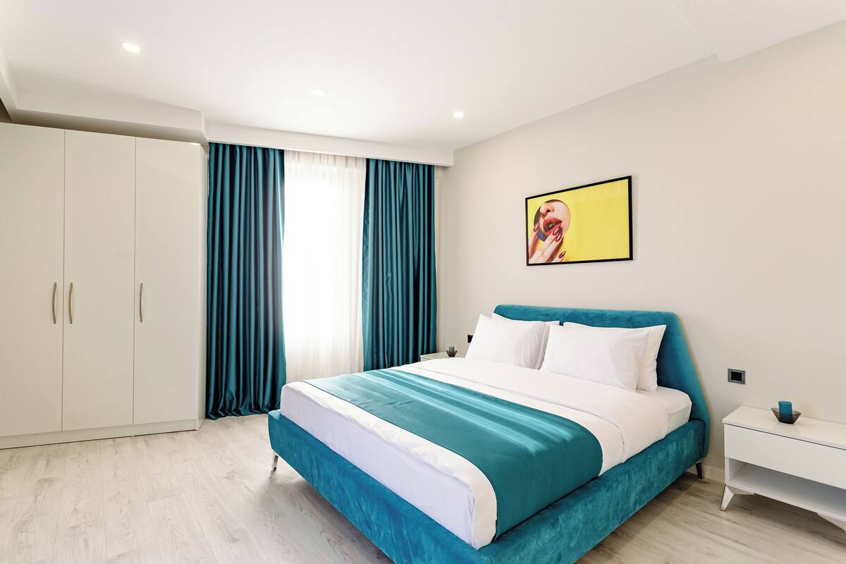 A modern bedroom with a large bed, white wardrobe, teal curtains, and a painting on the wall.