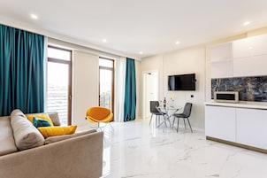 A modern living room with a sofa, a dining table, and chairs. There is a kitchen area with a microwave and cabinets.