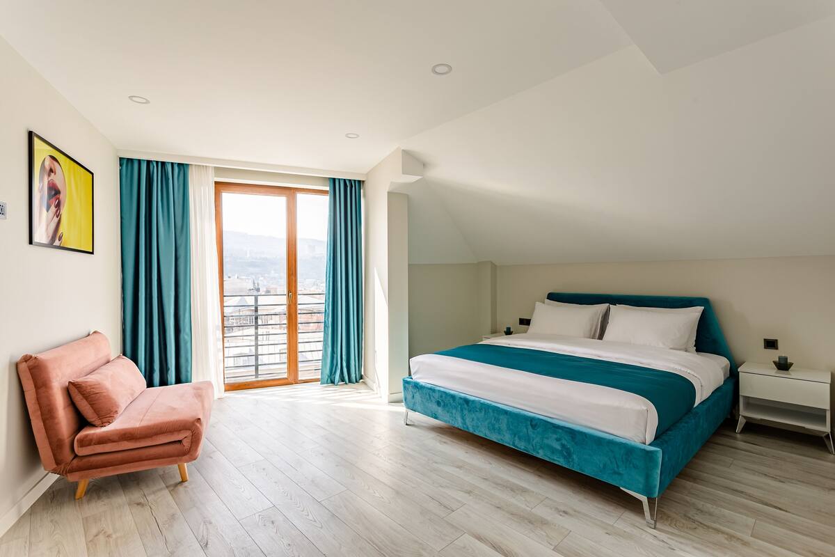 A bedroom with a bed, a chair, a nightstand, and a large window with curtains.