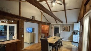 Ein geräumiger Essbereich mit hoher Decke und freiliegenden Holzbalken, eine Küche mit weißen Schränken und ein Blick ins Freie durch ein Fenster.