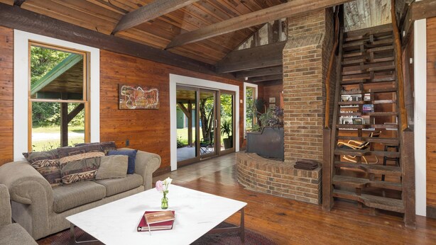 A spacious living area with a wooden ceiling, a brick fireplace, a built-in bookshelf, and a large window offering a view of the outdoors. - Cabin on the Crook: private, pet friendly residence alongside a nature preserve (Rising Fawn)