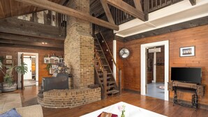 A spacious living room with a brick fireplace, wooden beams, a staircase, a television, and a dining table. - Cabin on the Crook: private, pet friendly residence alongside a nature preserve (Rising Fawn)