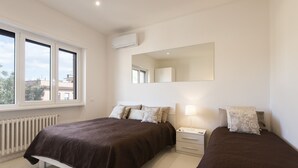 Ein Schlafzimmer mit zwei Betten, einem großen Fenster, einer Heizung, einem Nachttisch mit Lampe und einem Spiegel an der Wand.