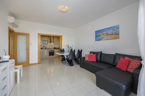 A modern living room with a black leather sofa, a dining table with chairs, a kitchen area, and a painting on the wall.