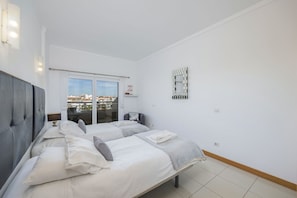 A bedroom with two beds, a view of the city through a sliding door, a wall-mounted mirror, and a small shelf with a decorative item.