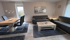 Een moderne woonkamer met een eethoek, grijze banken, een bijzettafeltje en een schilderij aan de muur.