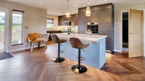 Une cuisine moderne dotée d’un îlot central, de deux tabourets de bar, d’un éclairage suspendu et offrant une vue sur l’extérieur grâce à une porte coulissante.