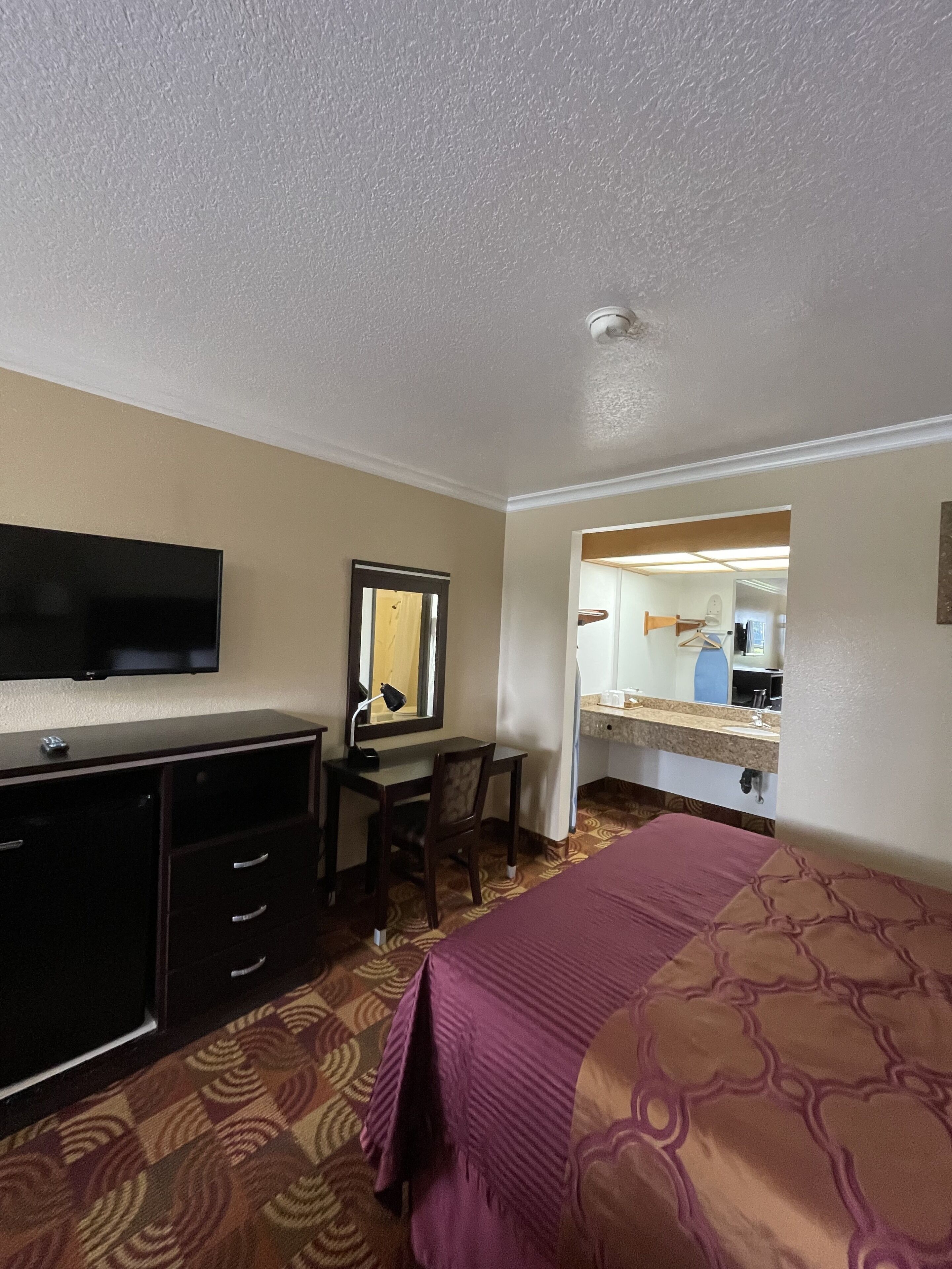 A hotel room with a bed, a desk with a chair, a television, a mirror, and a bathroom visible through an open door.