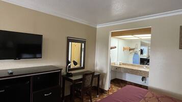 A hotel room with a bed, a desk with a chair, a television, a mirror, and a bathroom visible through an open door.